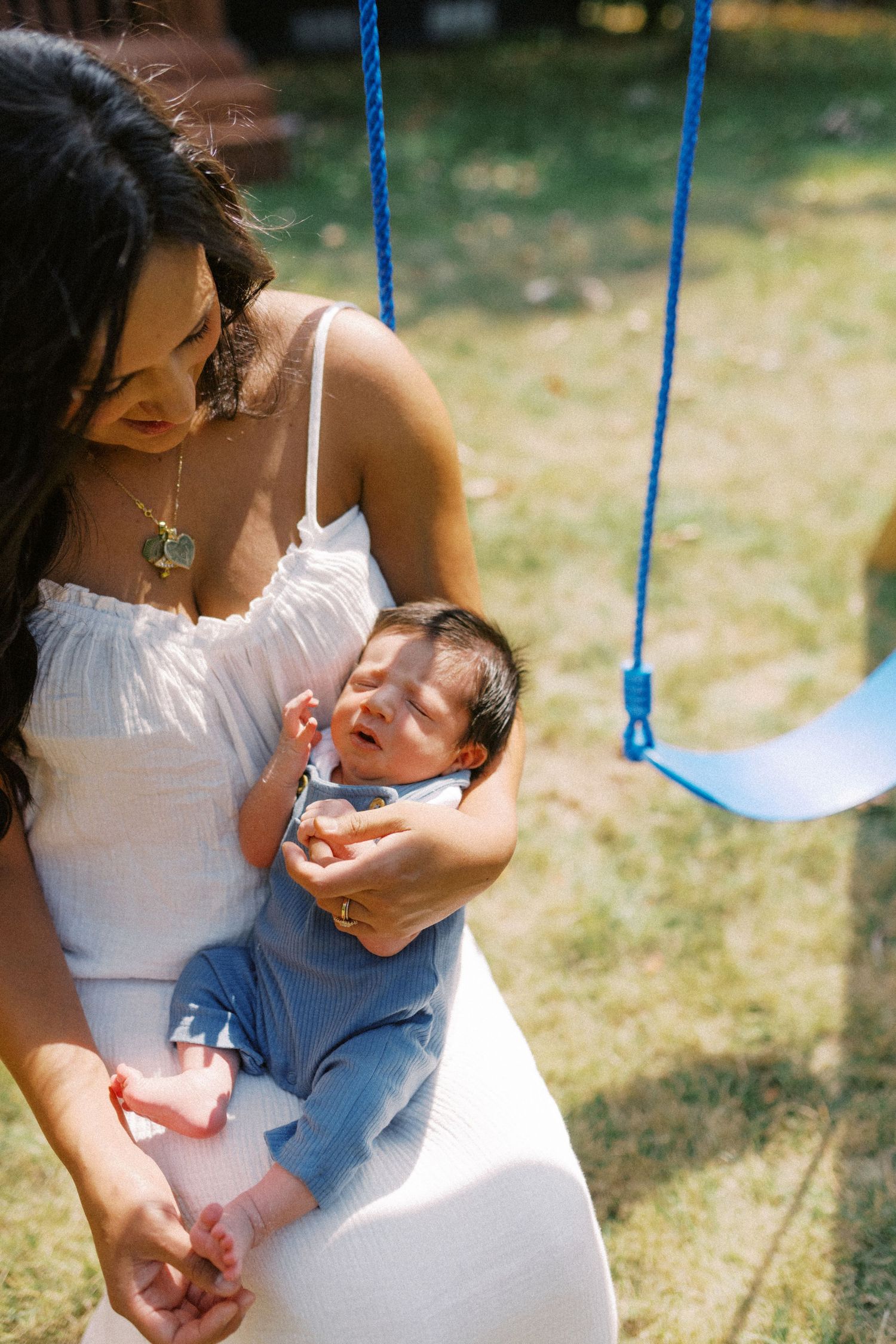 Mother in white dress cradling sleeping newborn baby in blue overalls outdoors near swing set.