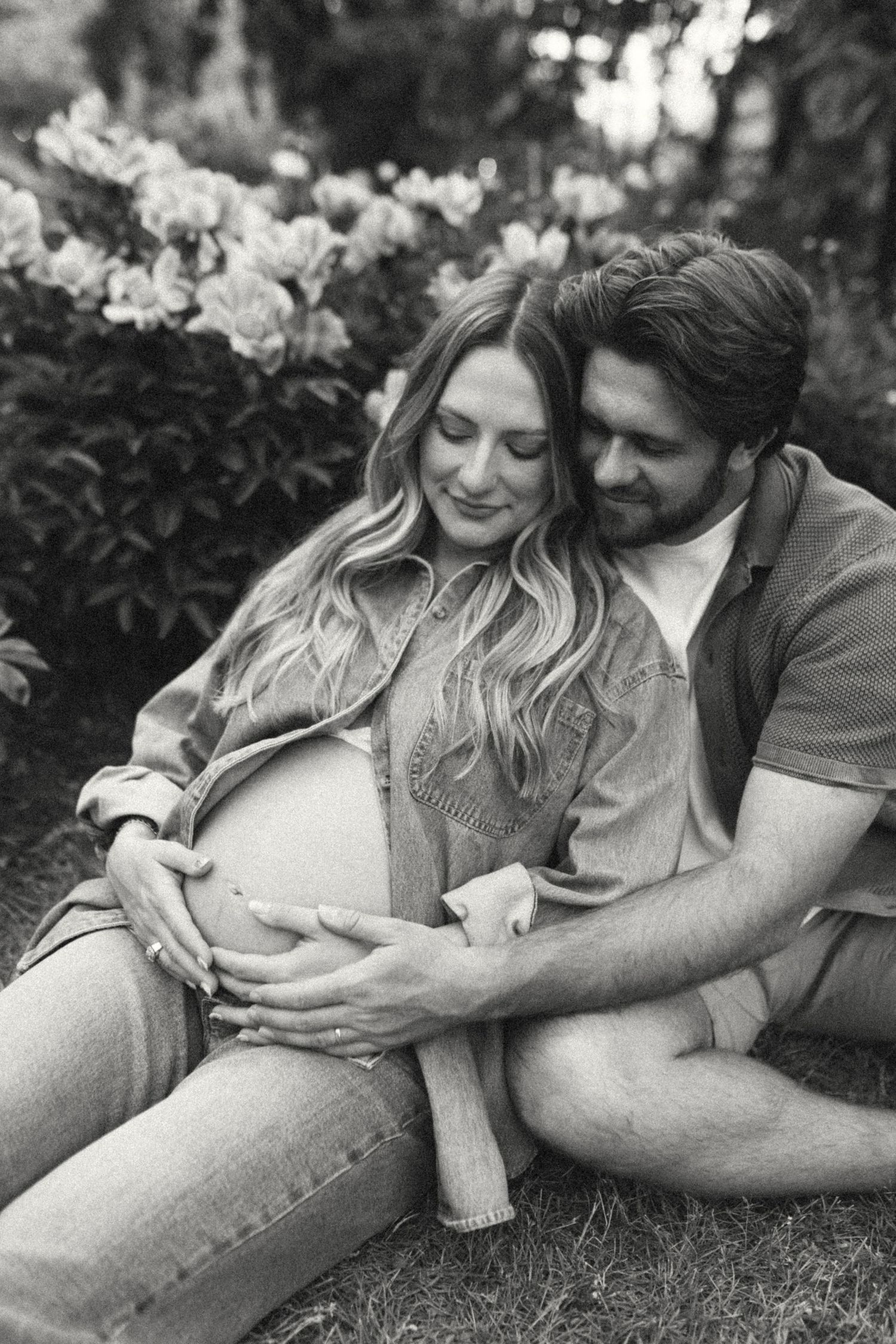 Black and white photo of couple embracing with hands on pregnant belly in garden.