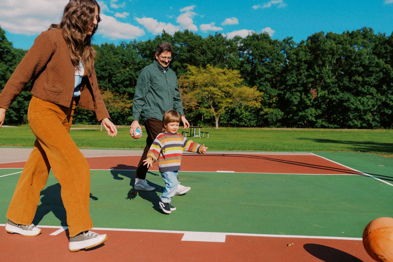 Family playing basketball outdoors on a court, with two adults and a toddler enjoying a sunny day surrounded by green trees.
