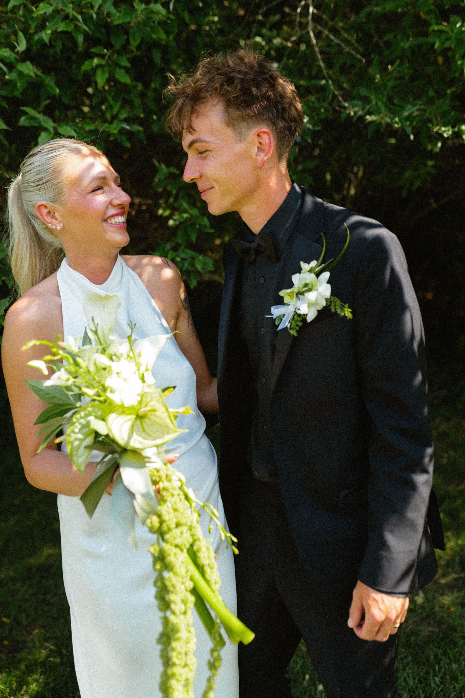Smiling bride in white halter dress with green bouquet and groom in black tuxedo.