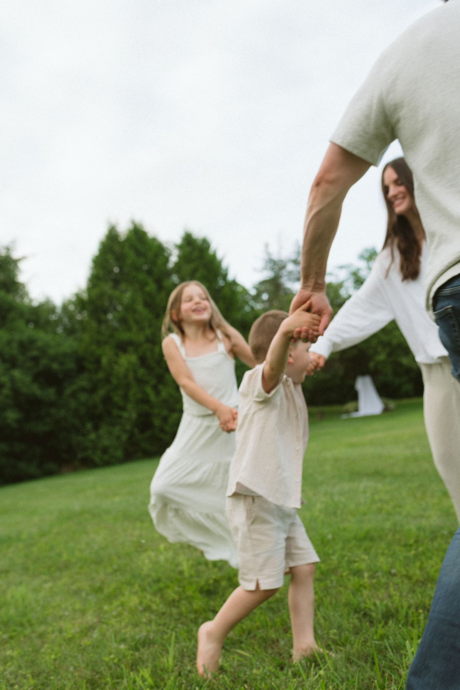 Family playing ring-around-the-rosy on green grass outdoors, children laughing in white outfits.