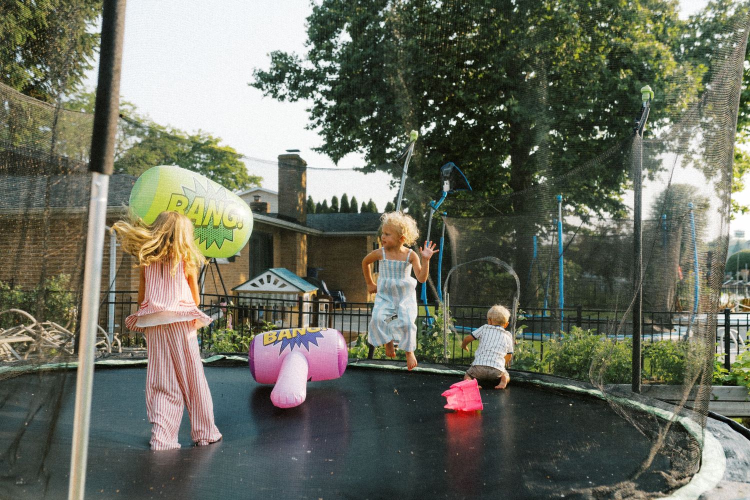 Three young children jumping on a backyard trampoline surrounded by green trees on a sunny day.