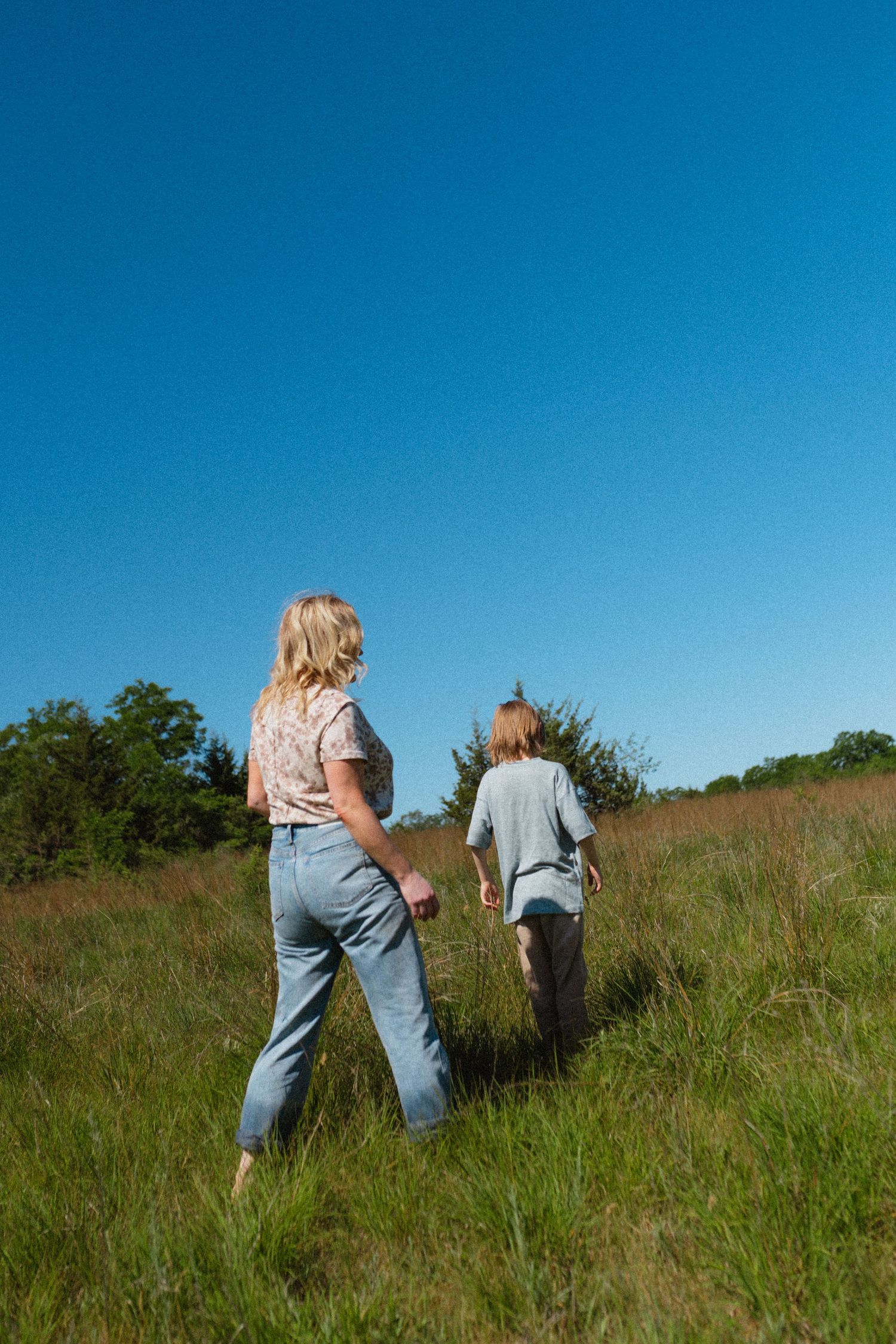 Mother and son walking barefoot through tall green grass field under clear blue sky.
