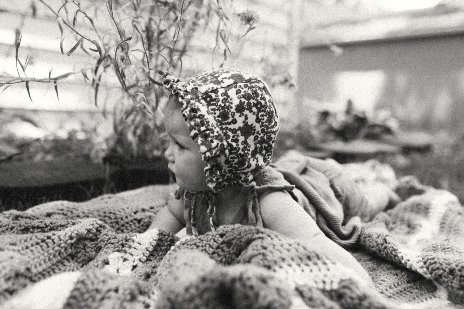 Black and white photo of a baby in a patterned bonnet lying on a crocheted blanket outdoors.