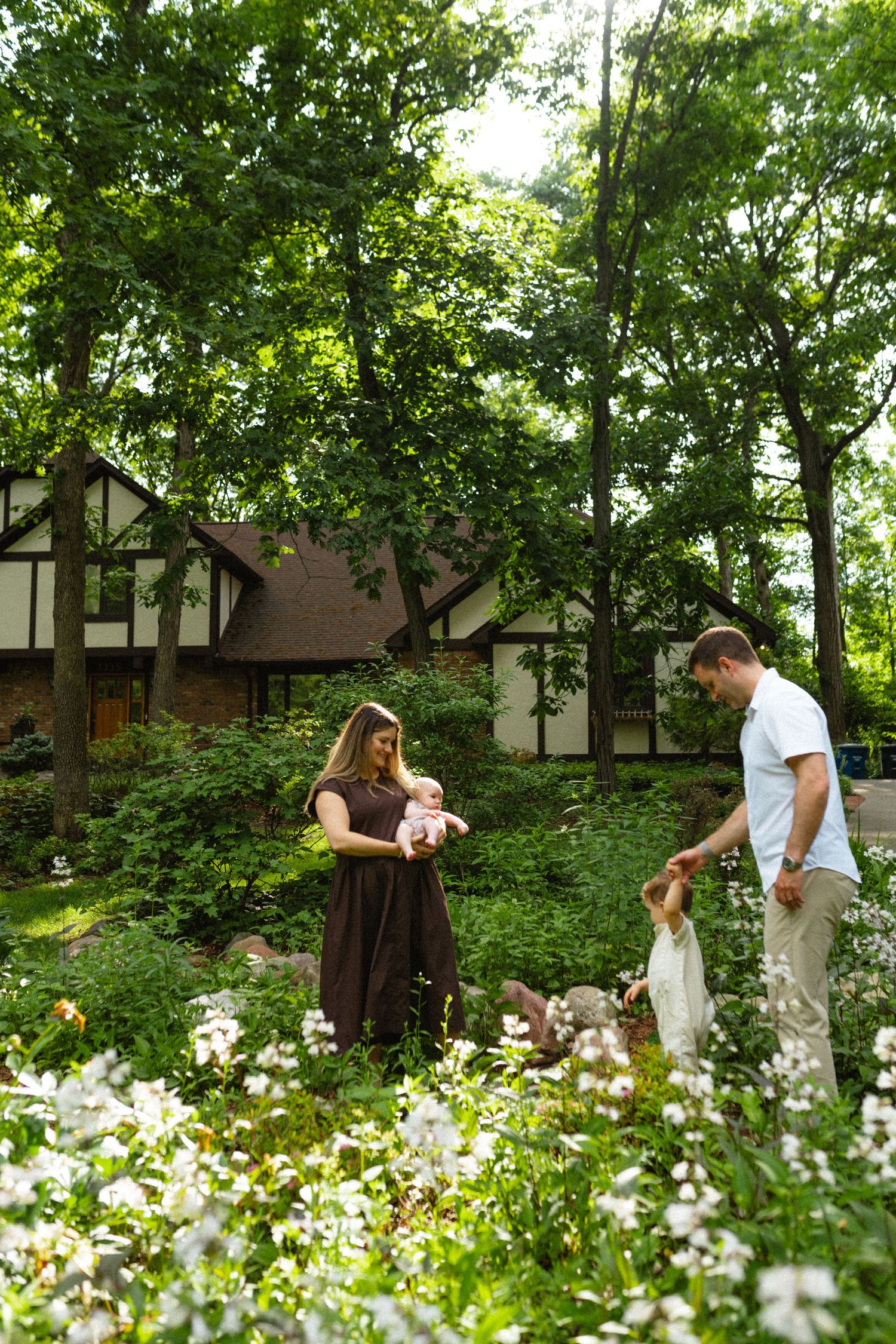 Family with baby and toddler exploring lush garden with white wildflowers outside Tudor home.