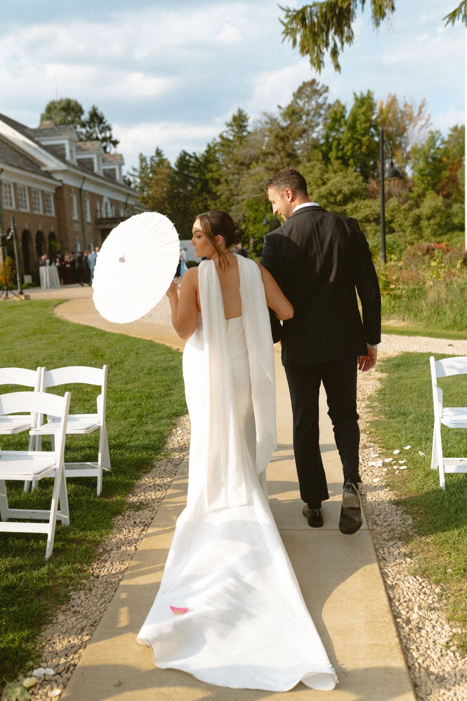 Bride in white gown holding parasol walks with groom in black suit along outdoor wedding aisle.