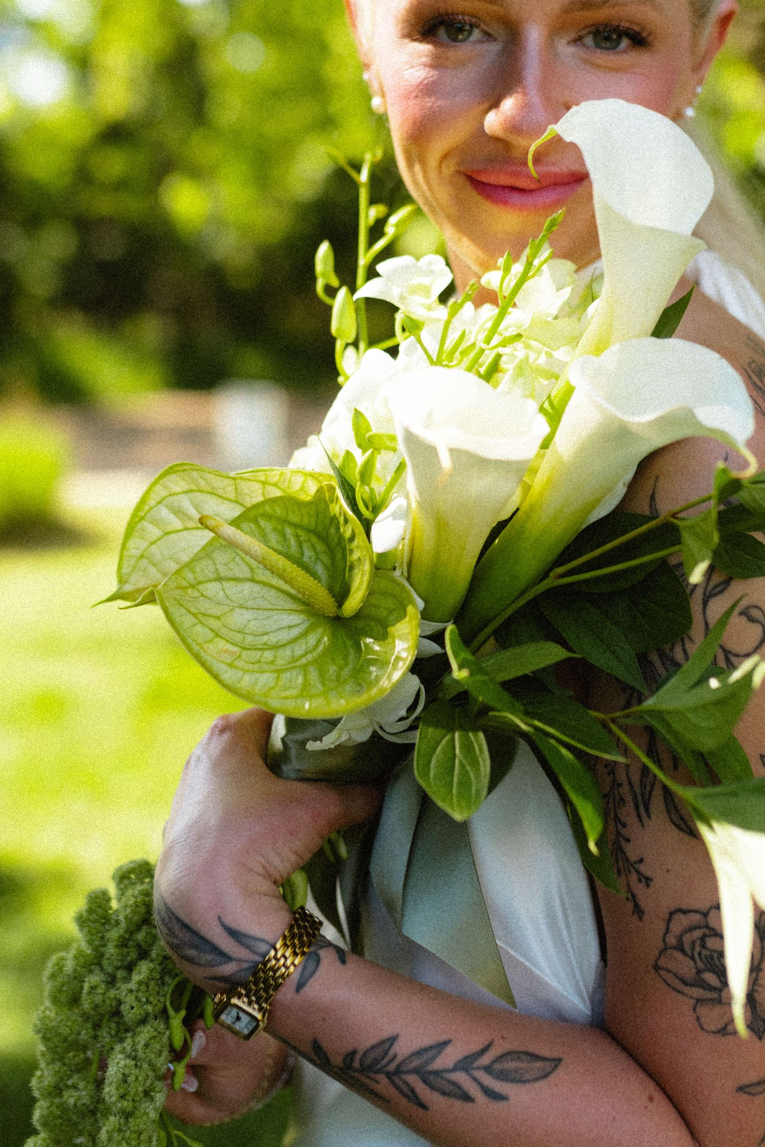 Bride with tattoos smelling white calla lily and anthurium wedding bouquet outdoors.