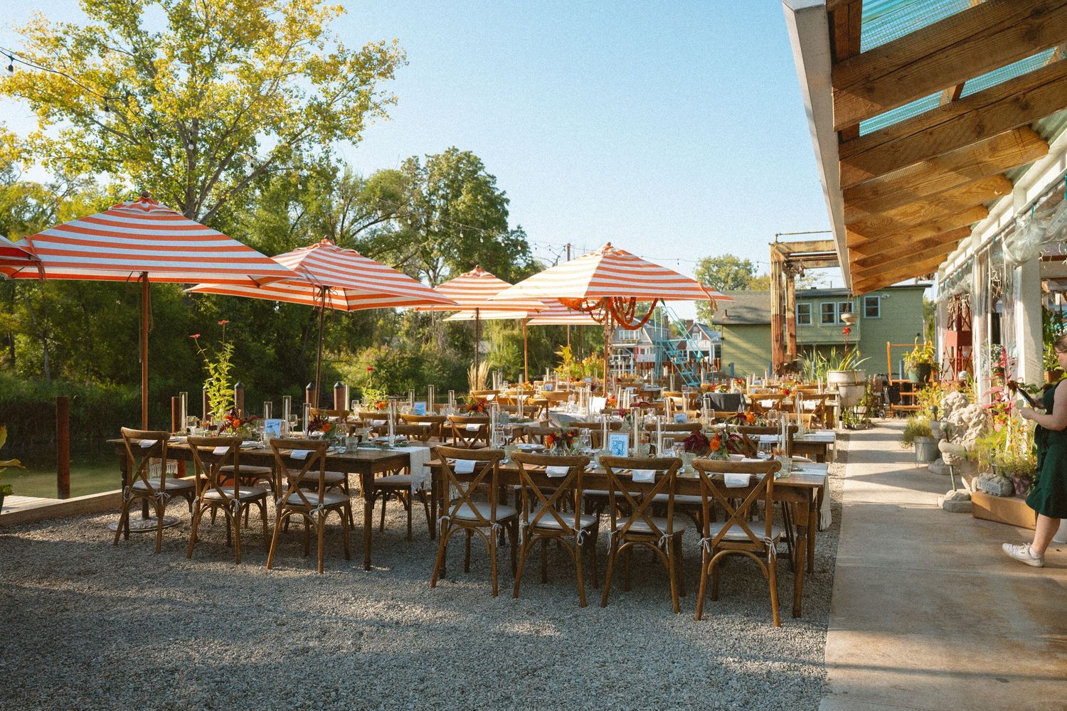 Outdoor wedding reception venue with striped red and white umbrellas, wooden tables and chairs on gravel.