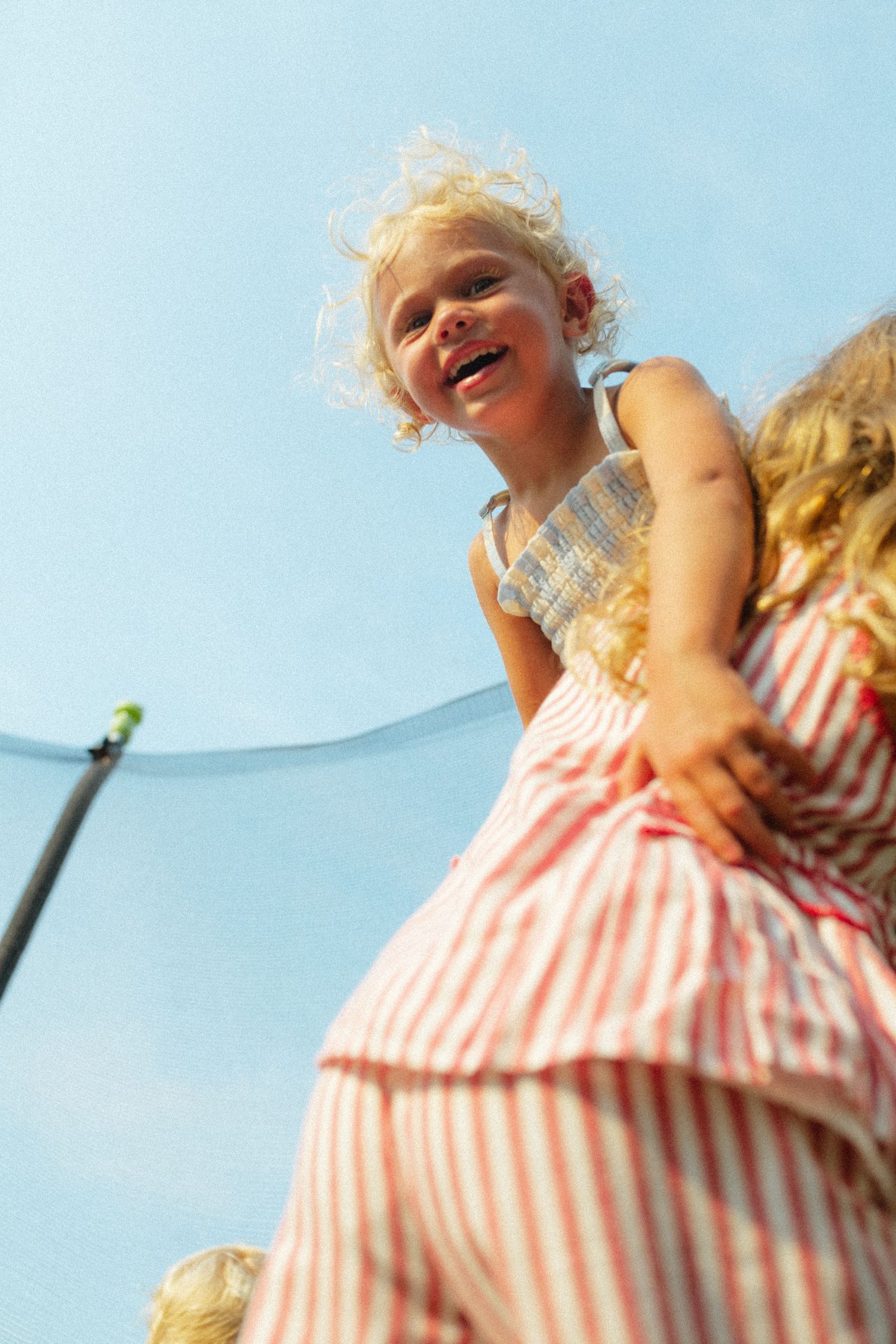 Happy blonde toddler girl laughing joyfully on a trampoline against a bright blue sky.