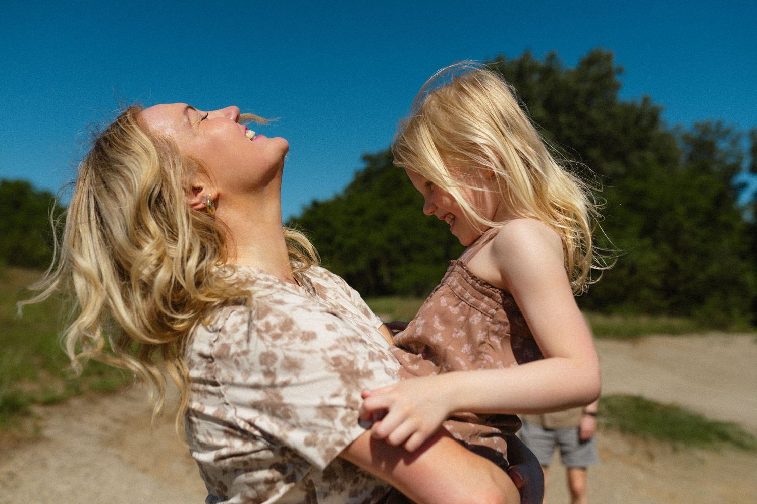 Mother laughing joyfully while holding blonde toddler daughter outdoors on sunny day.