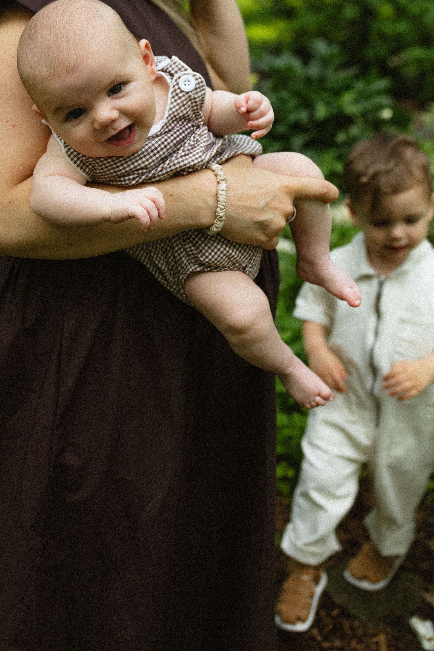 Smiling baby in brown gingham romper held by mother outdoors with toddler sibling in white jumpsuit nearby.