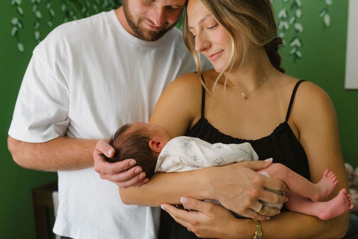 Couple tenderly gazing at their newborn baby wrapped in white blanket against green wall.