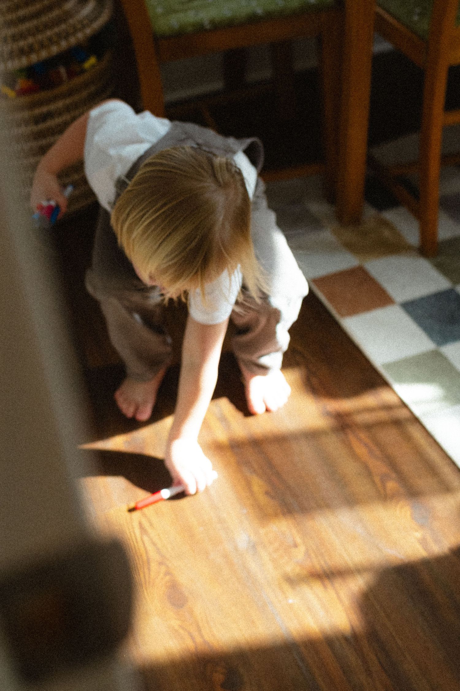 Young blonde child in overalls drawing on a sunlit hardwood floor.