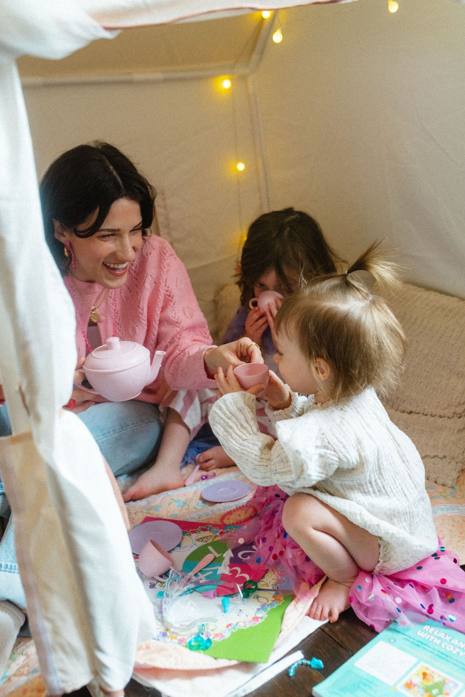 Mother and two young daughters having a tea party inside a cozy blanket fort with fairy lights.