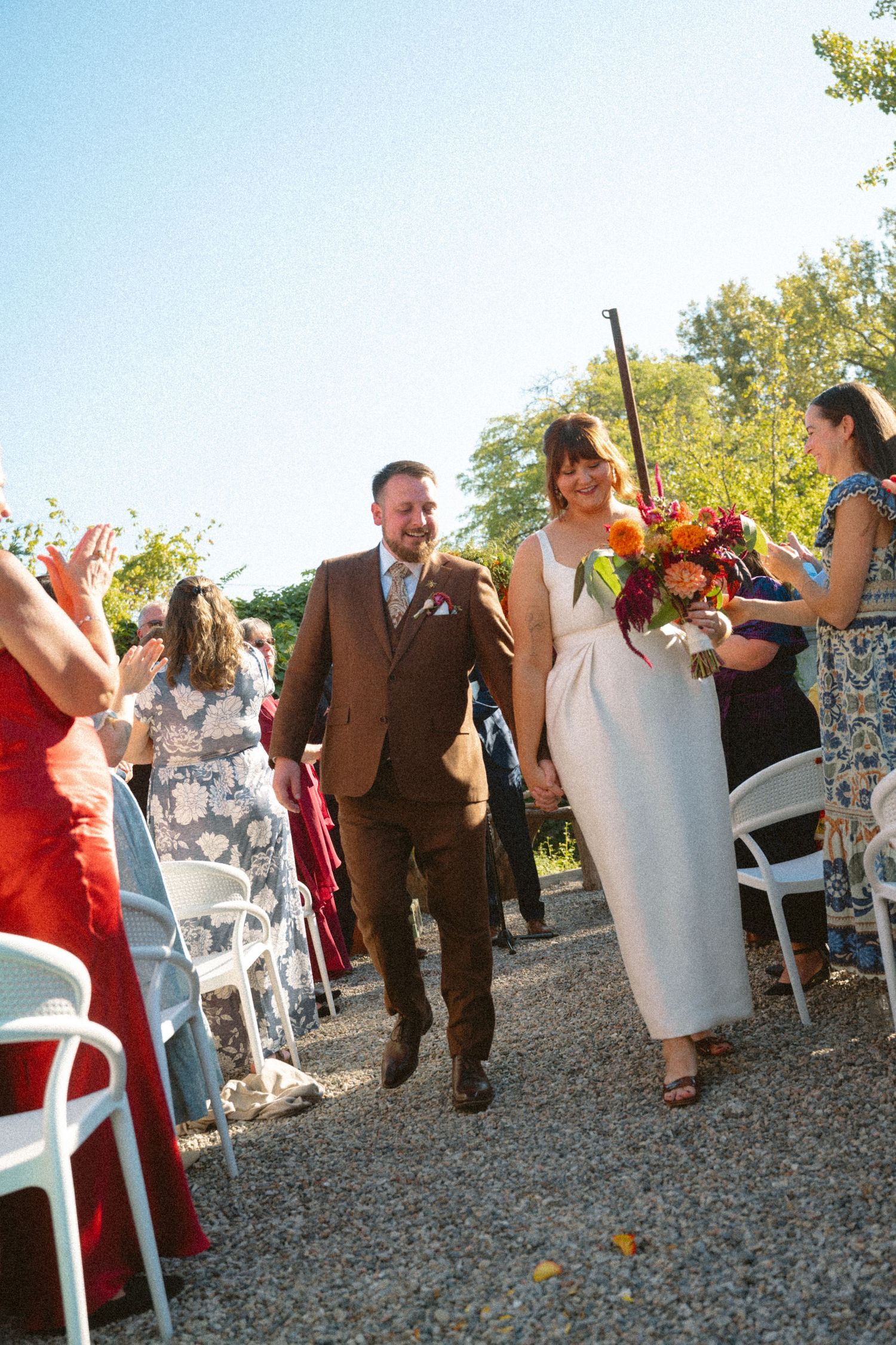 Joyful newlywed couple walking down outdoor aisle after wedding ceremony, bride holding vibrant orange floral bouquet.