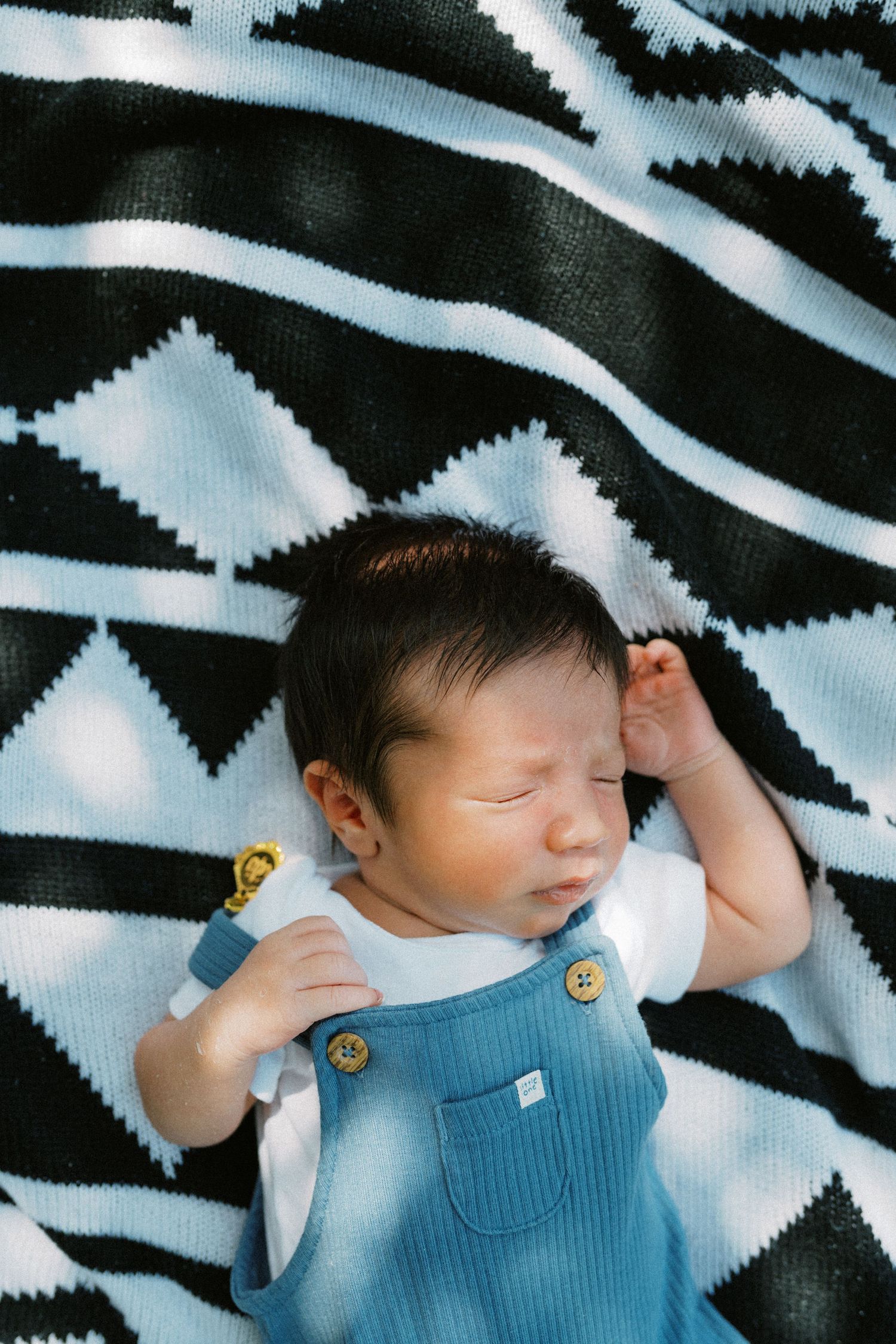 Peaceful newborn baby sleeping in blue overalls on a black and white patterned blanket outdoors.