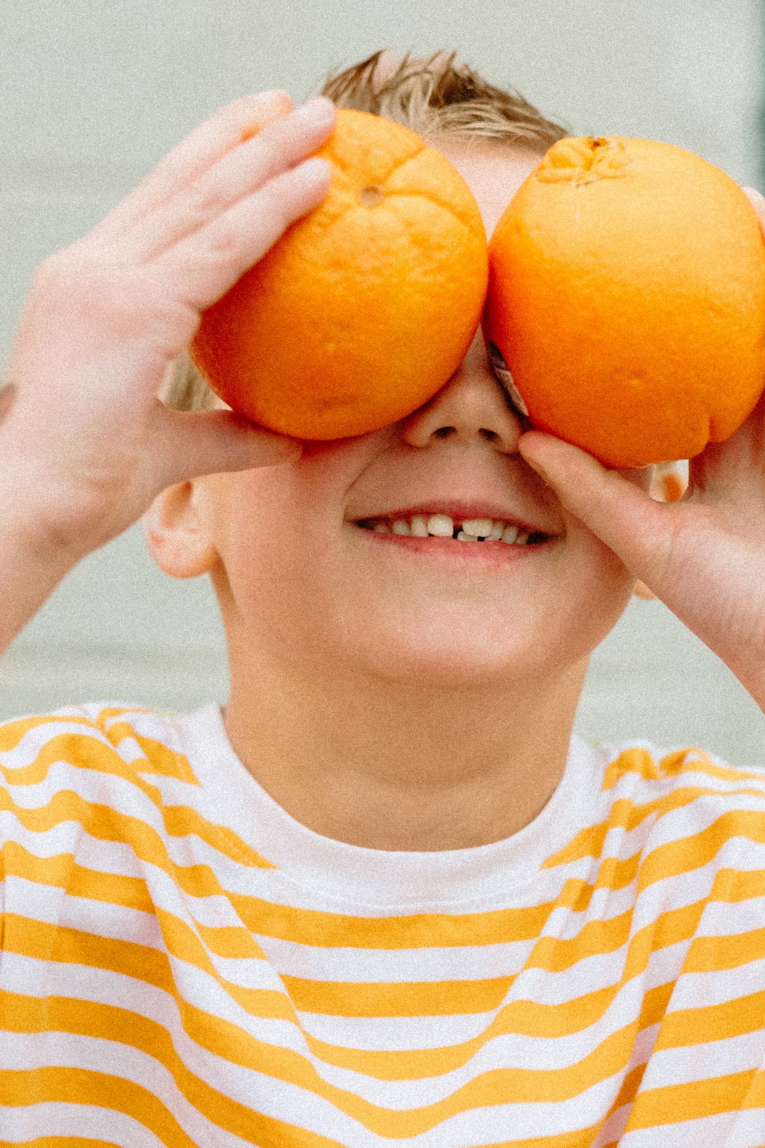 Happy boy in yellow striped shirt holding two oranges over his eyes smiling.