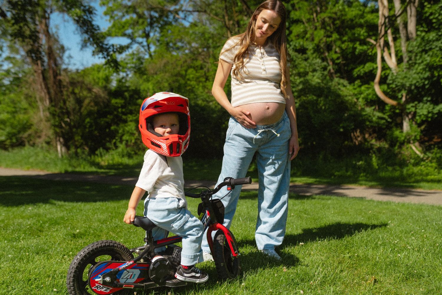 Pregnant mother in jeans watches young child in red helmet ride a small bike on green grass in a sunny park.