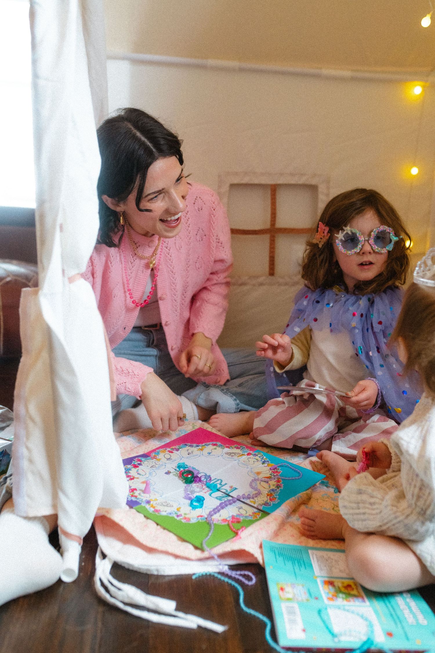 Woman in pink cardigan laughing while playing craft games with young girl wearing novelty glasses indoors.