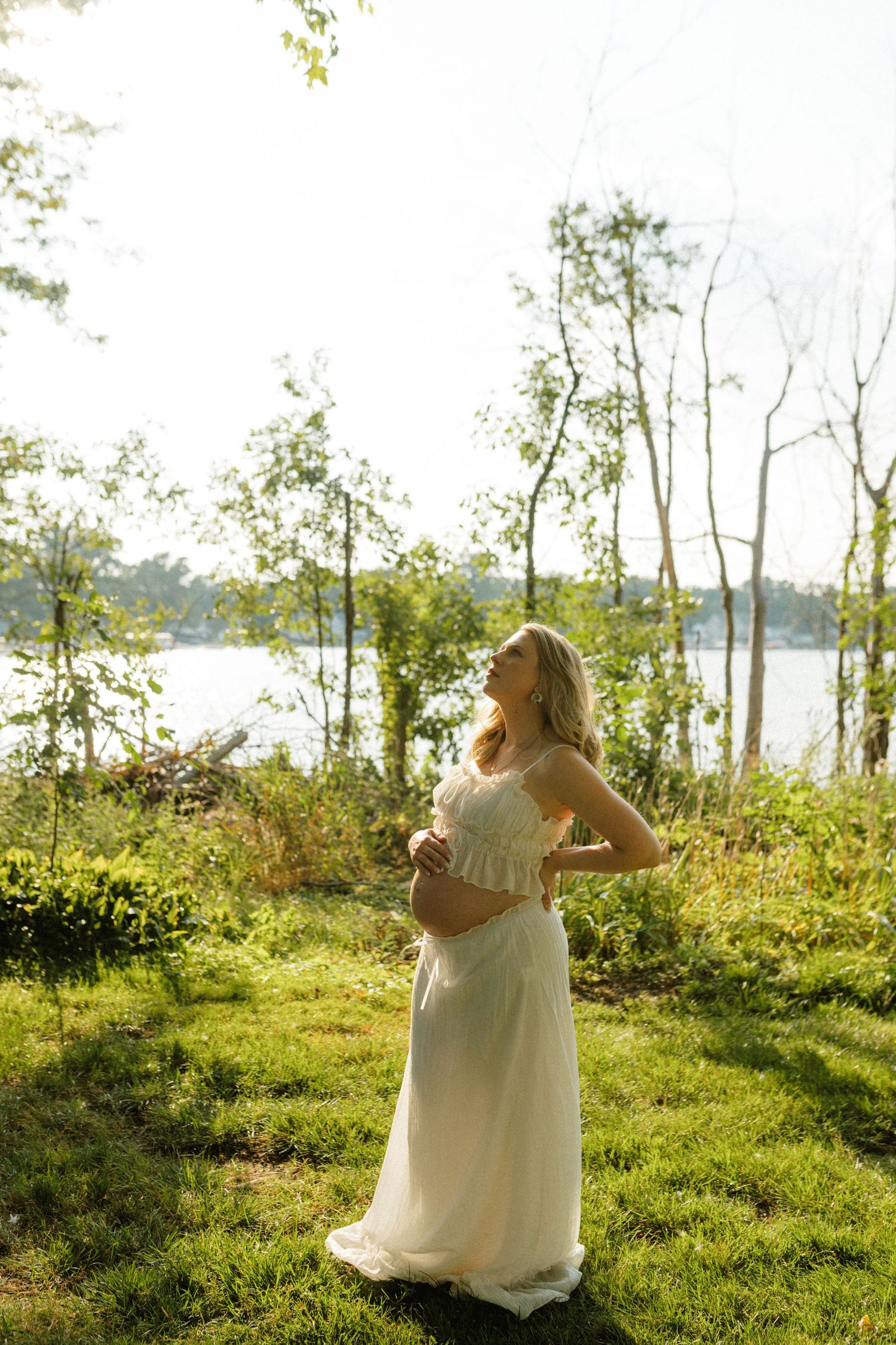 A pregnant woman in a white dress standing outdoors near a lake, looking up in golden sunlight.