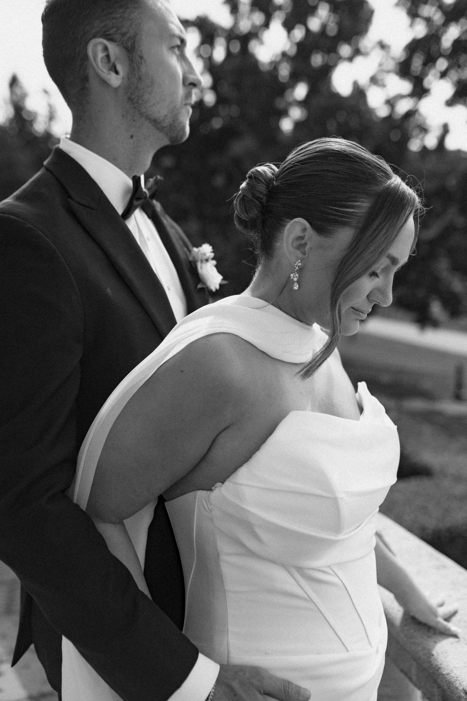 Black and white portrait of groom embracing bride from behind in elegant wedding attire.