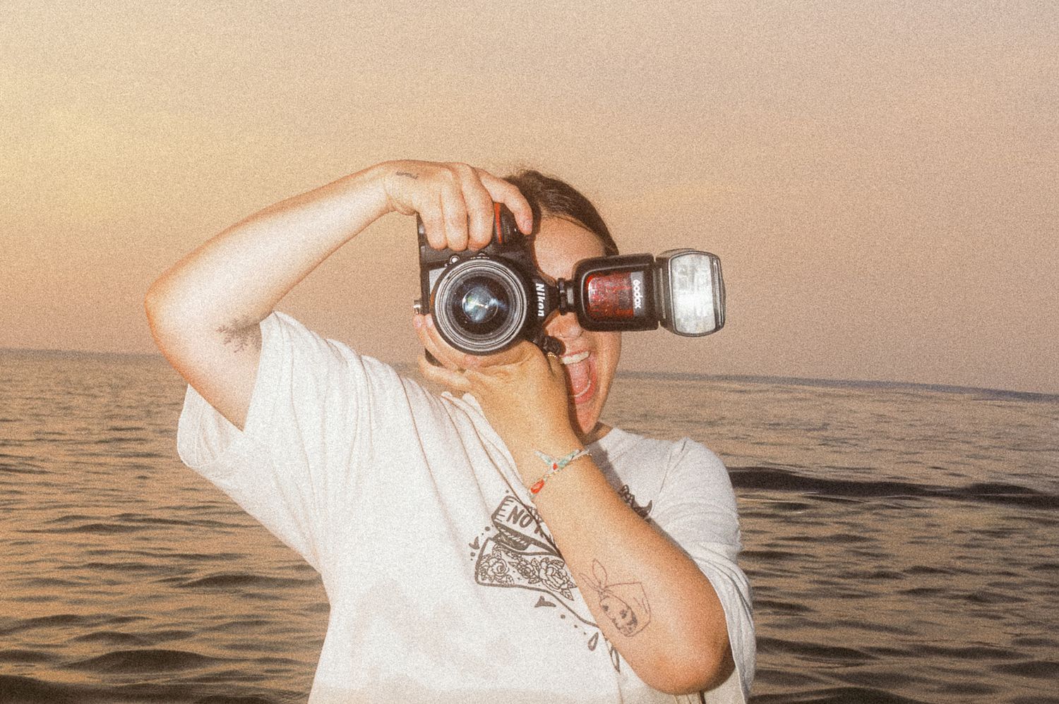Photographer holding a film camera with flash up to their face while standing near the ocean at sunset.