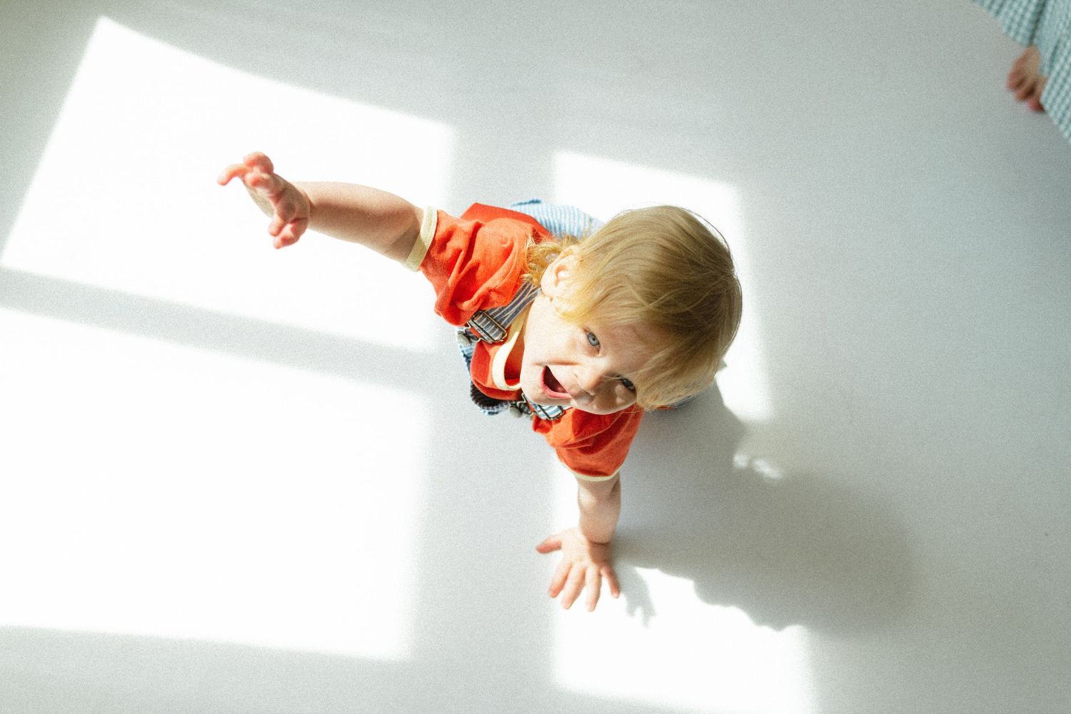 Overhead view of a toddler in orange shirt reaching out on a bright white floor.