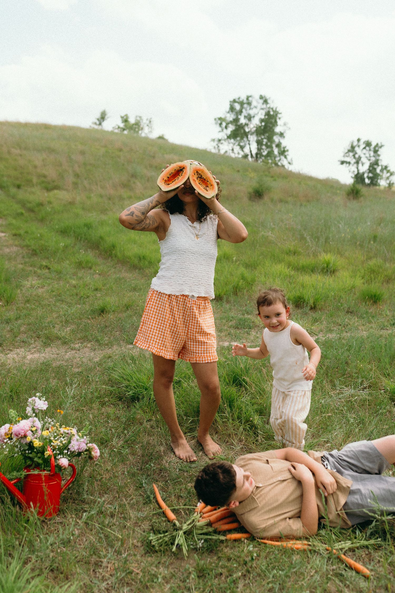 Mother holds cantaloupe halves over eyes while children laugh during outdoor family picnic.