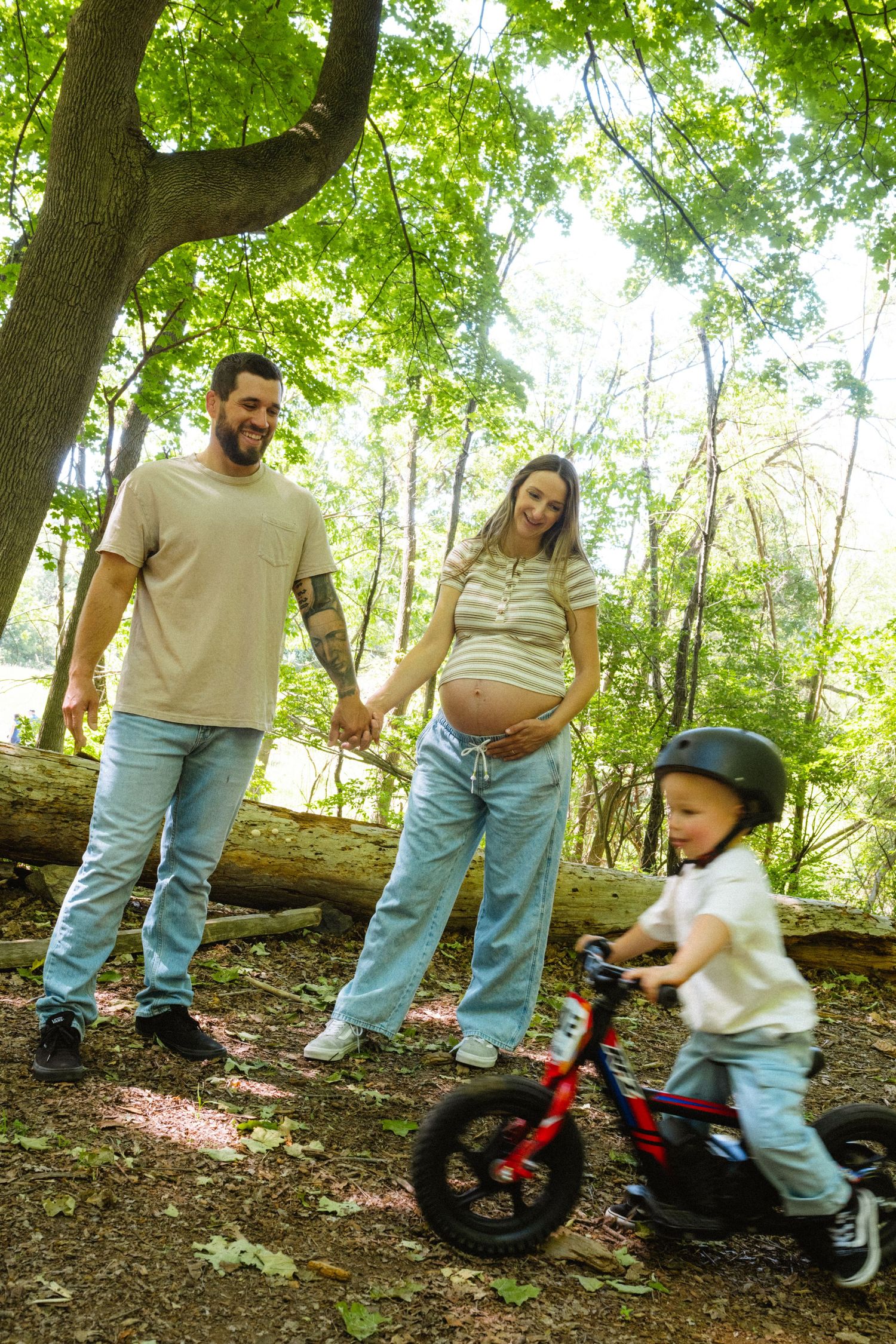 Expecting couple holding hands watching toddler ride balance bike in lush green forest park.