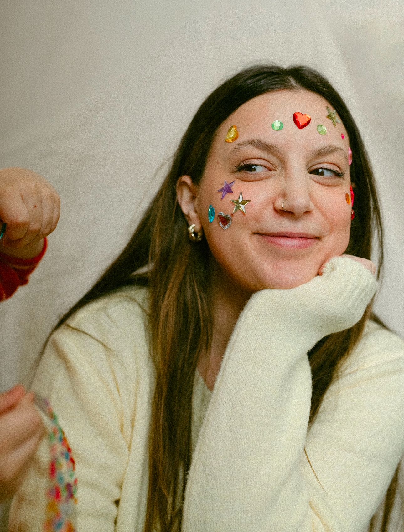 Young woman with colorful gem stickers on face smiles warmly wearing a white sweater.