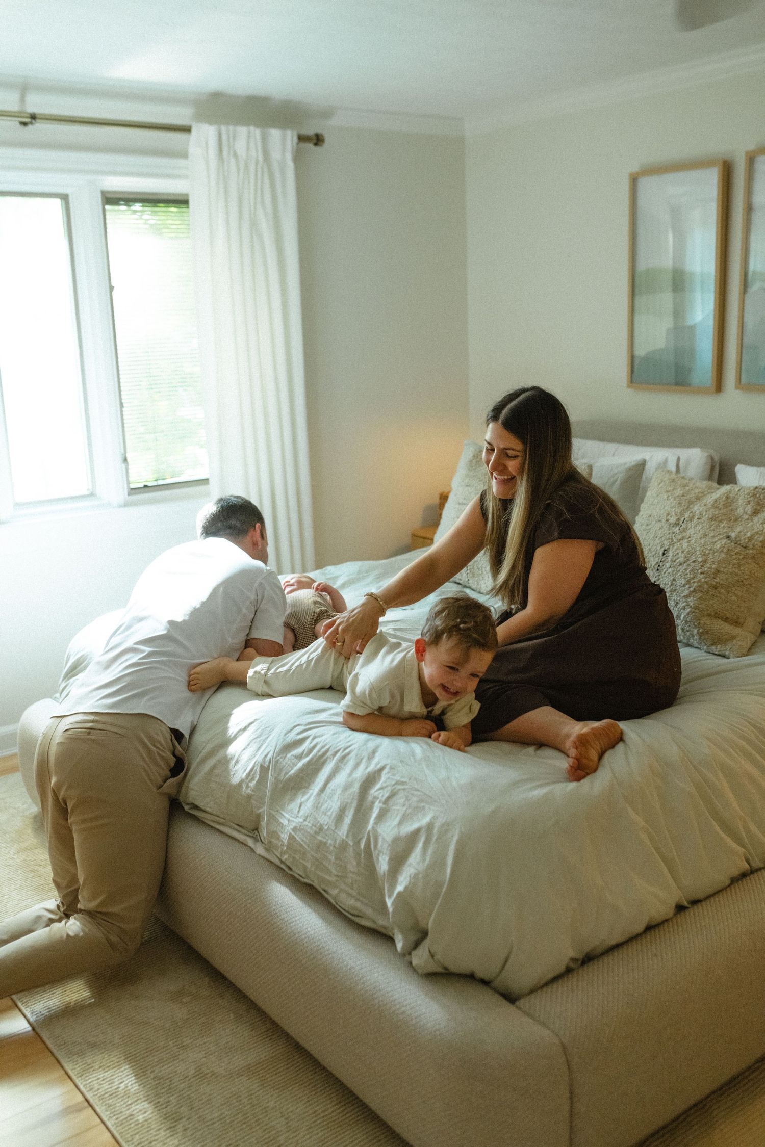 Parents playfully interacting with toddler and newborn on a large white bed in a bright bedroom.