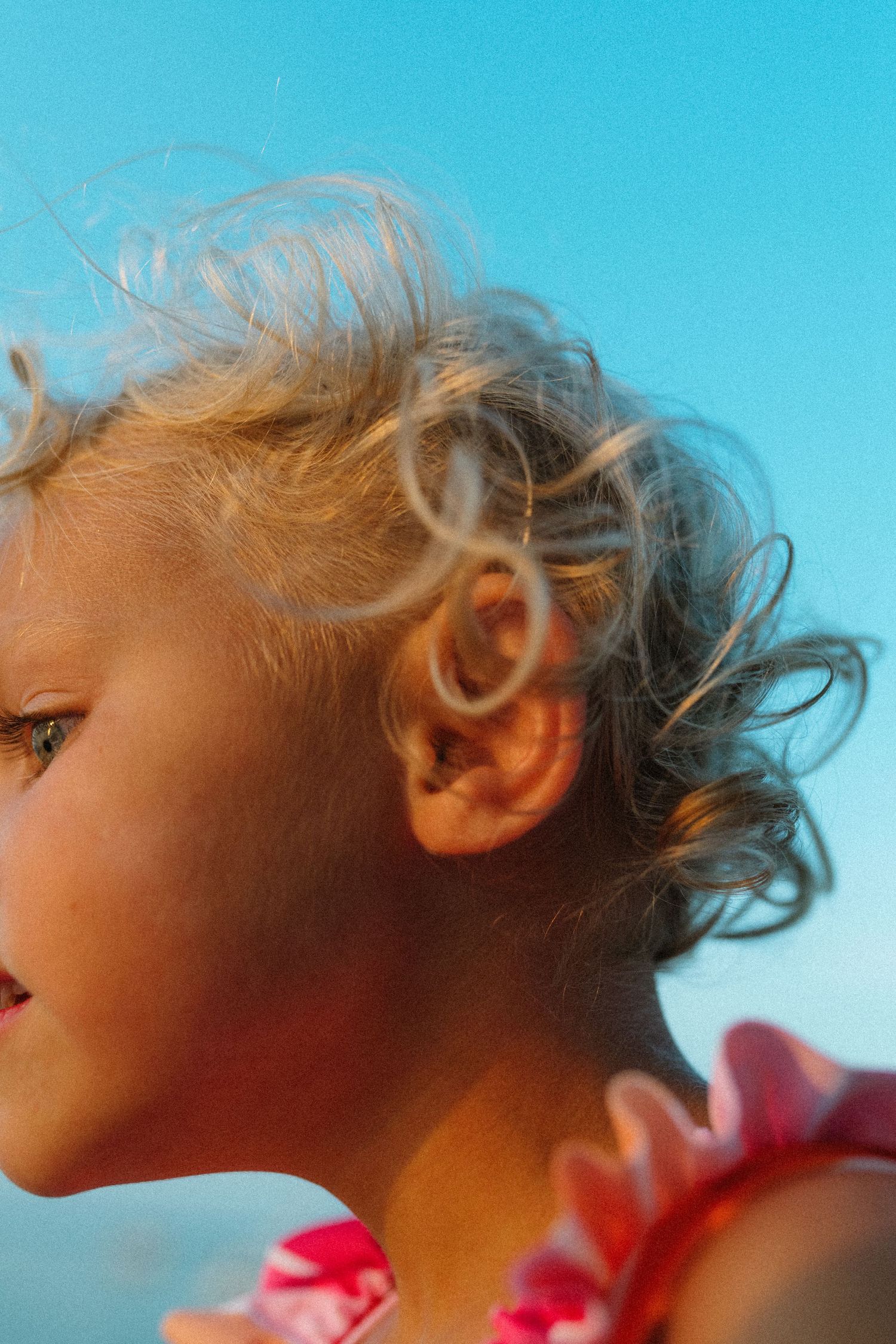 Close-up profile portrait of a young blonde child with curly hair against a blue sky.