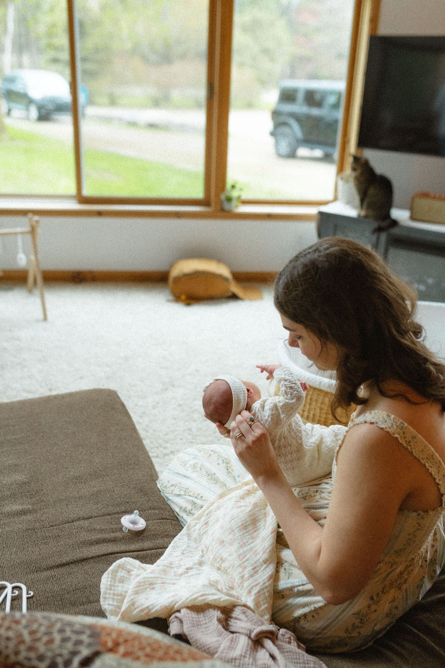 New mother sitting on floor cradling newborn baby in sunlit living room with cat.