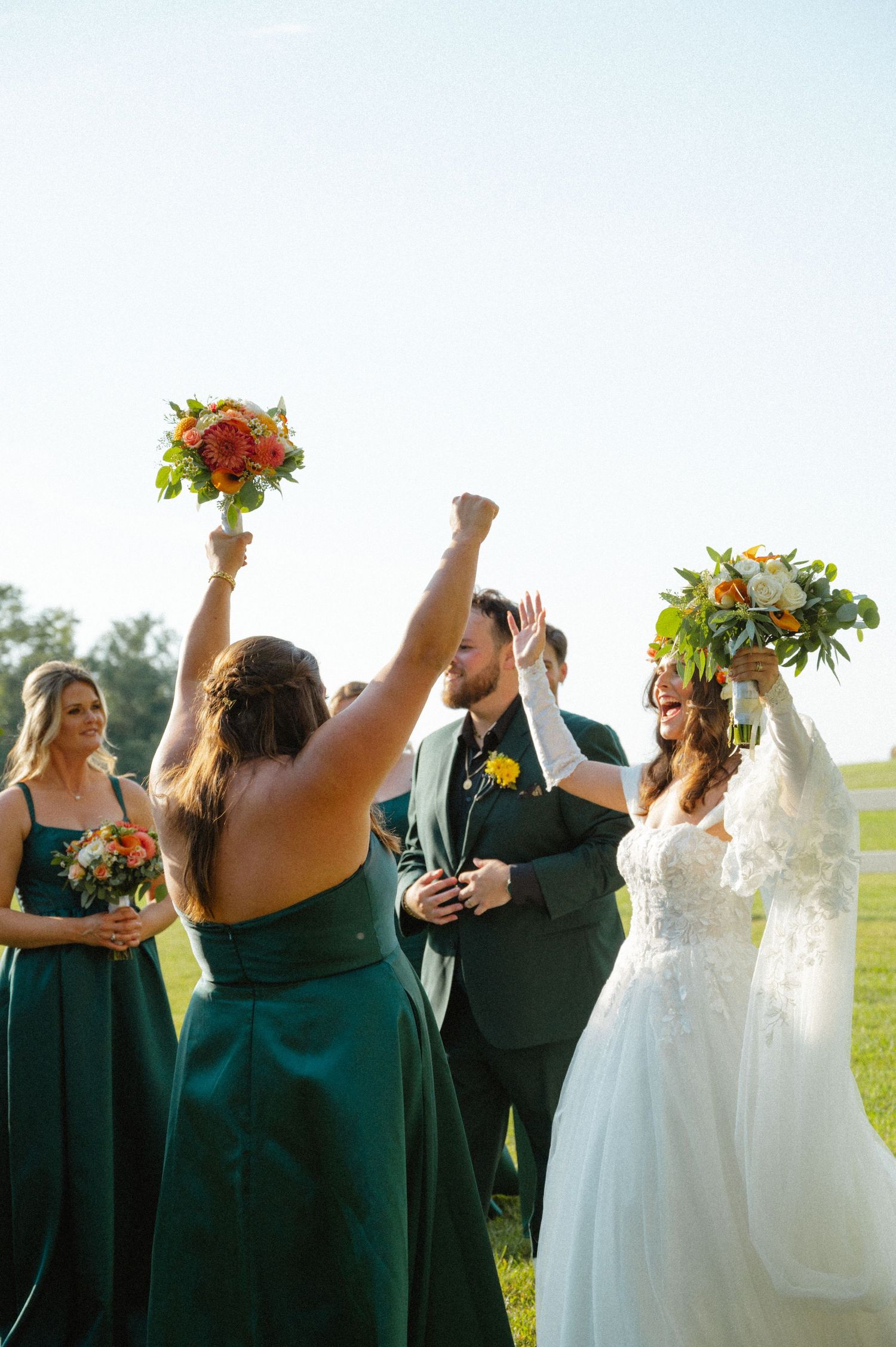 Wedding party celebrating outdoors with bridesmaids in emerald dresses raising bouquets joyfully.