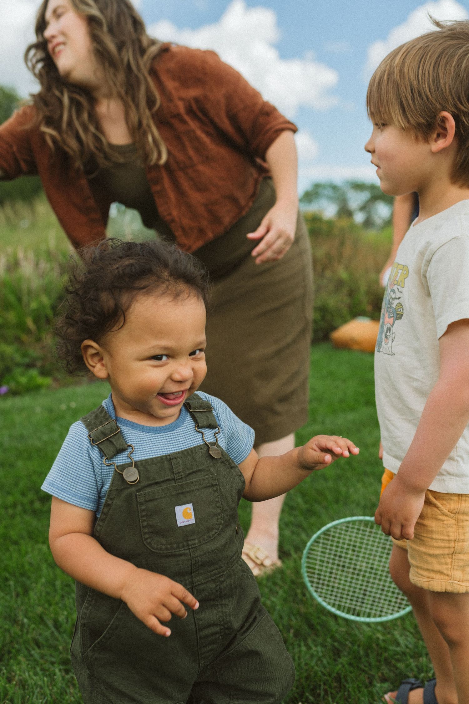 Toddler in overalls smiling outdoors while playing badminton with family in lush green yard.