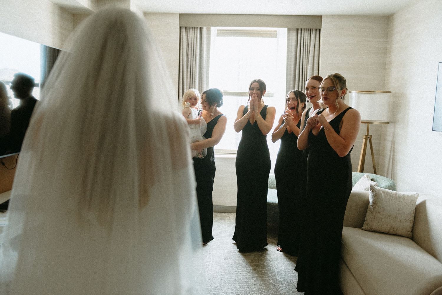 Bridesmaids in black dresses react emotionally during first look at bride in hotel room.