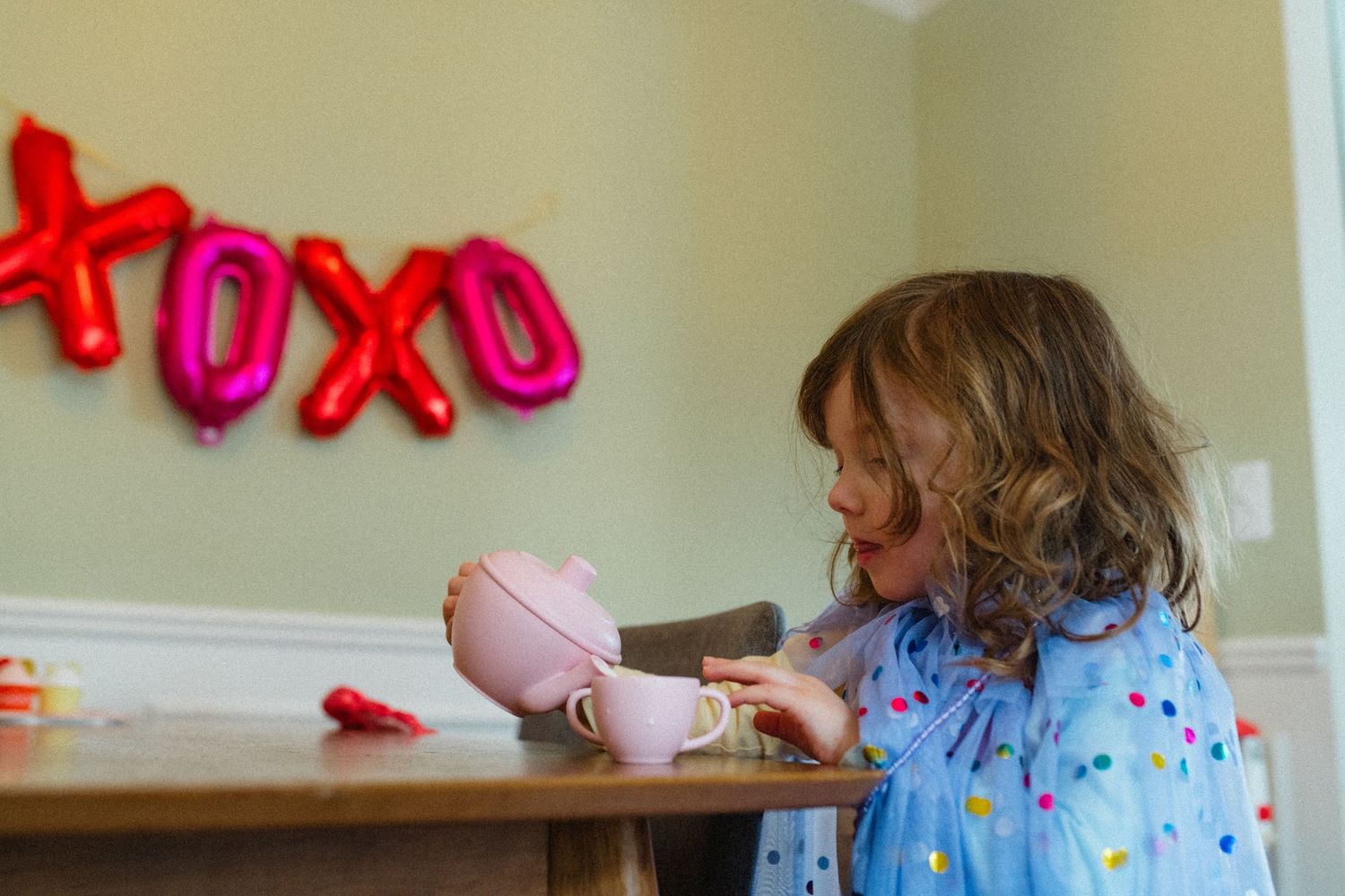 Young girl in blue polka dot dress playing with pink toy tea set near XOXO Valentine balloons.