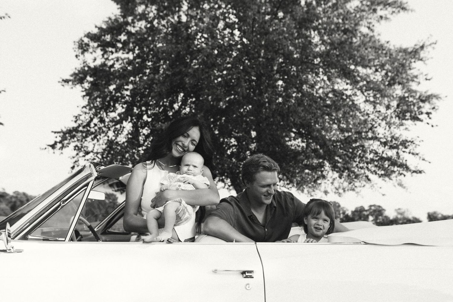 Black and white photo of family relaxing on hood of a classic convertible car.