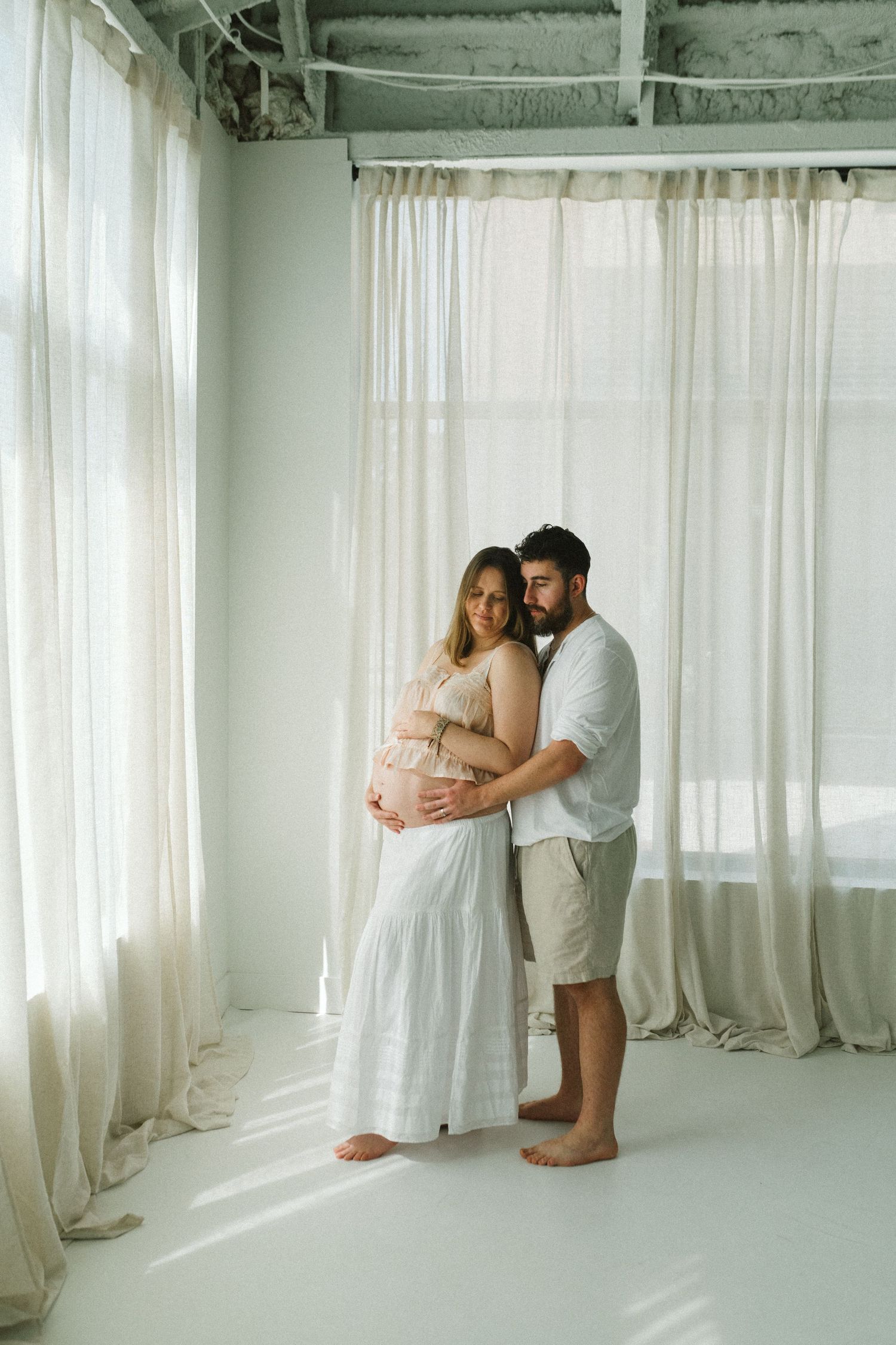Pregnant couple embracing tenderly in a bright white studio with sheer curtains in background.