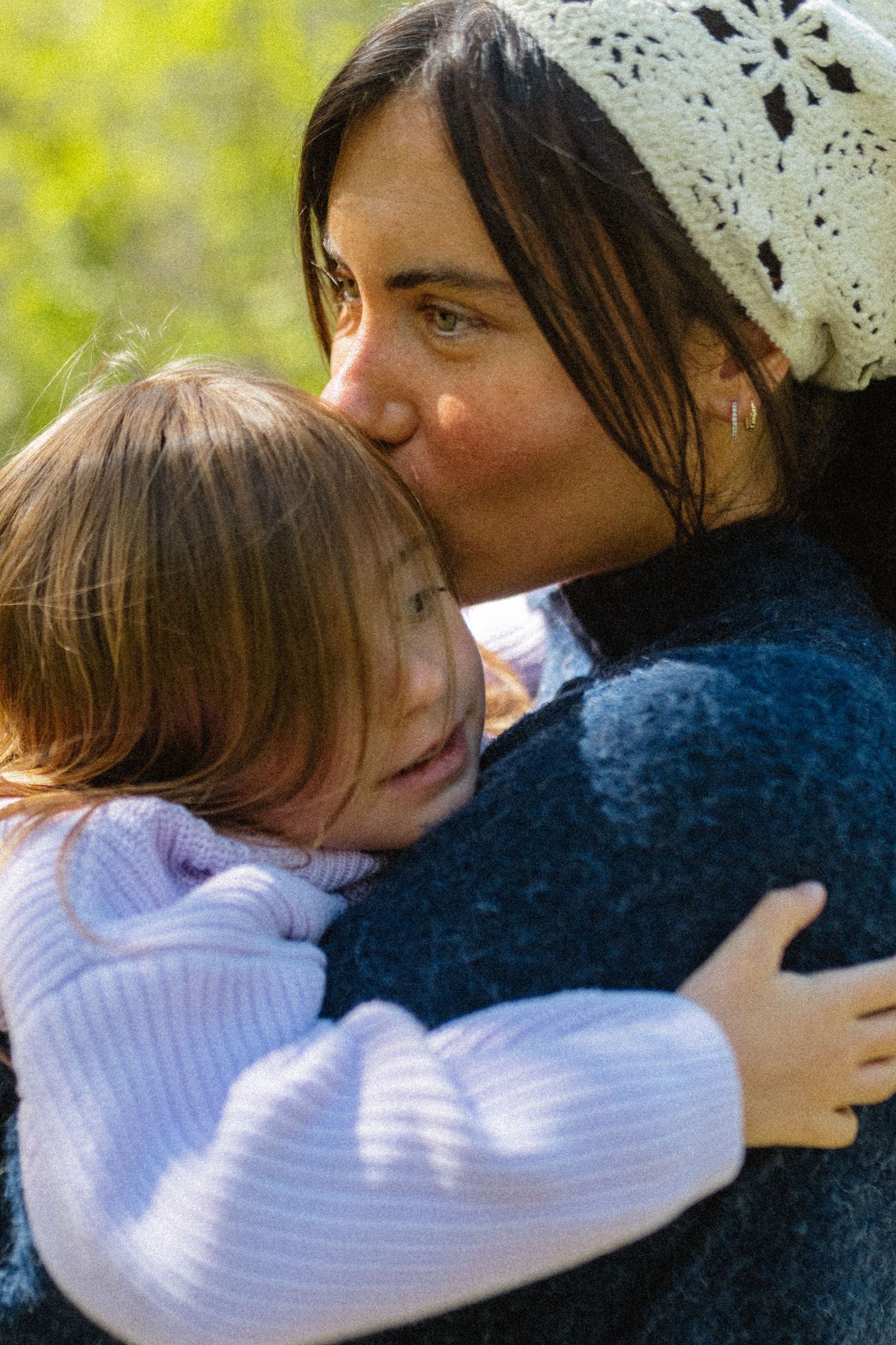 Mother in blue sweater tenderly kissing her young daughter on the head outdoors in nature.