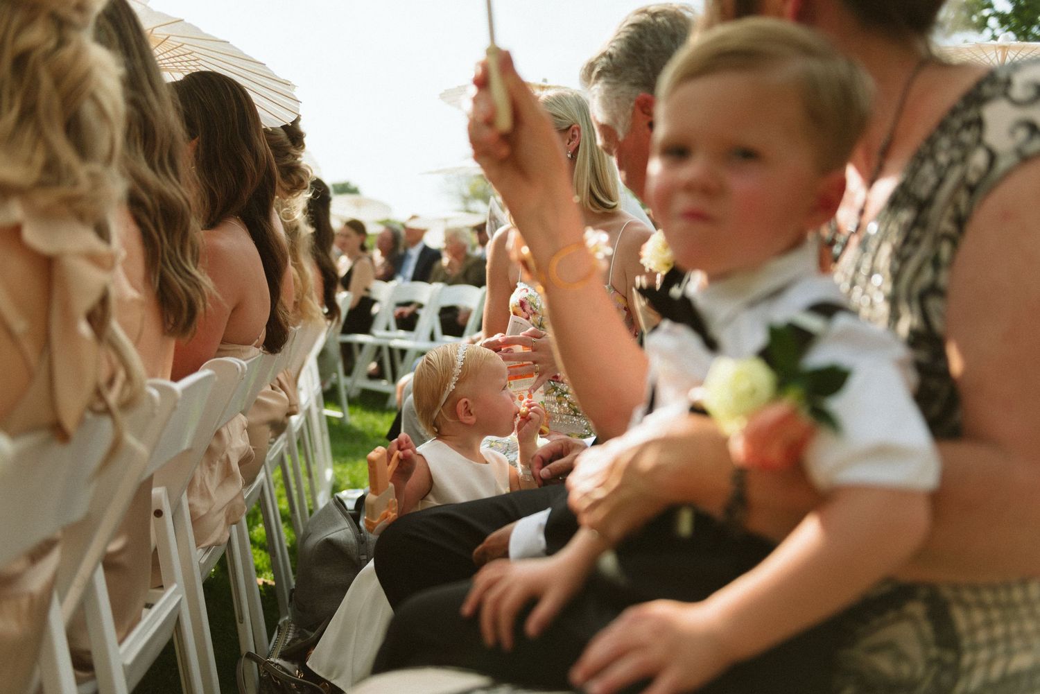 Wedding guests seated in white chairs watching outdoor ceremony with children in foreground.