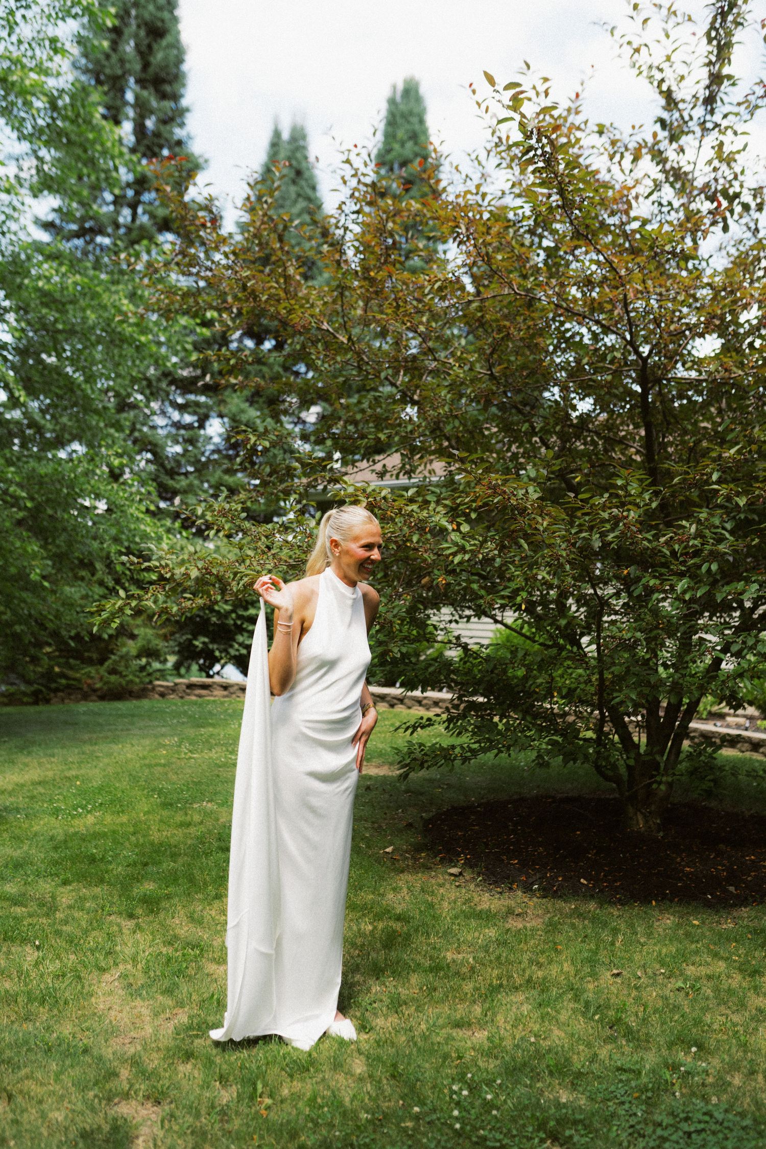 Bride in elegant white halter gown standing in lush green garden on wedding day.