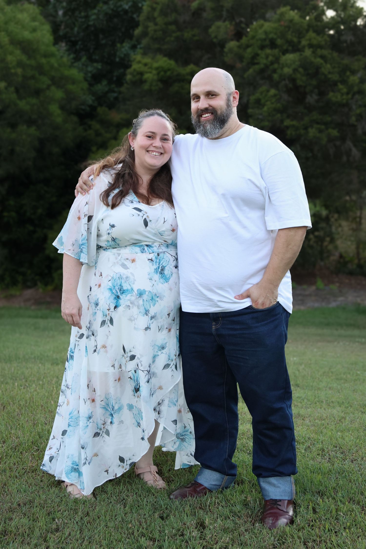 A smiling couple poses outdoors on green grass, woman in floral dress, bald bearded man in white tee and jeans.