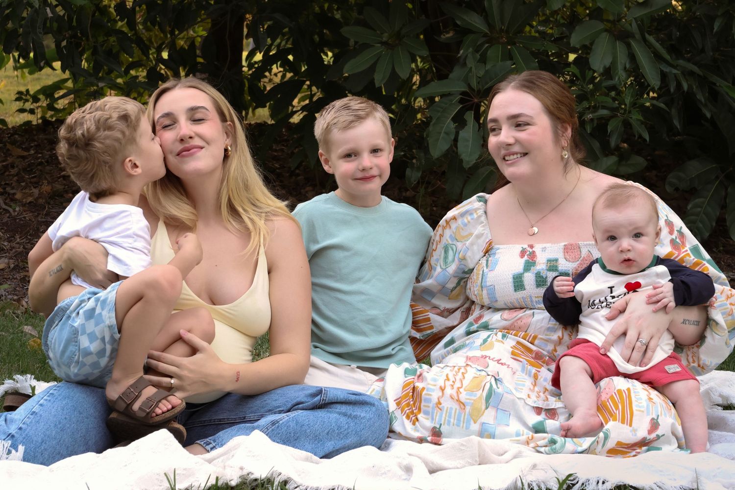 Two women sitting on a blanket outdoors with three young children, smiling together in a lush green garden setting.
