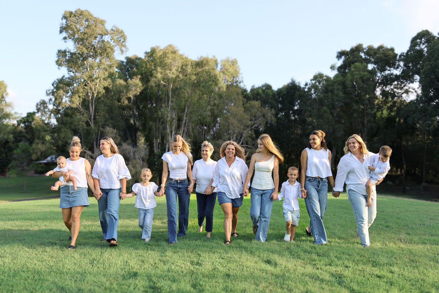 Group of women and children in white tops and jeans walking together on green grass park with trees.