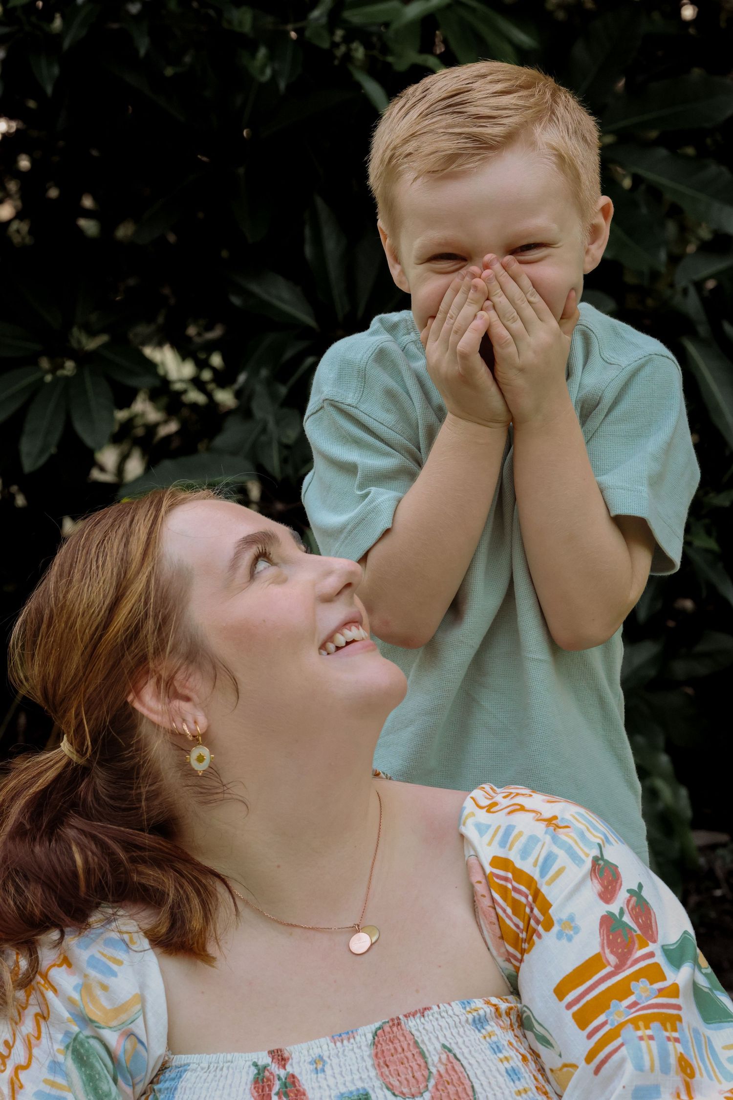 A laughing redhead woman looks up at a giggling young boy covering his mouth outdoors.