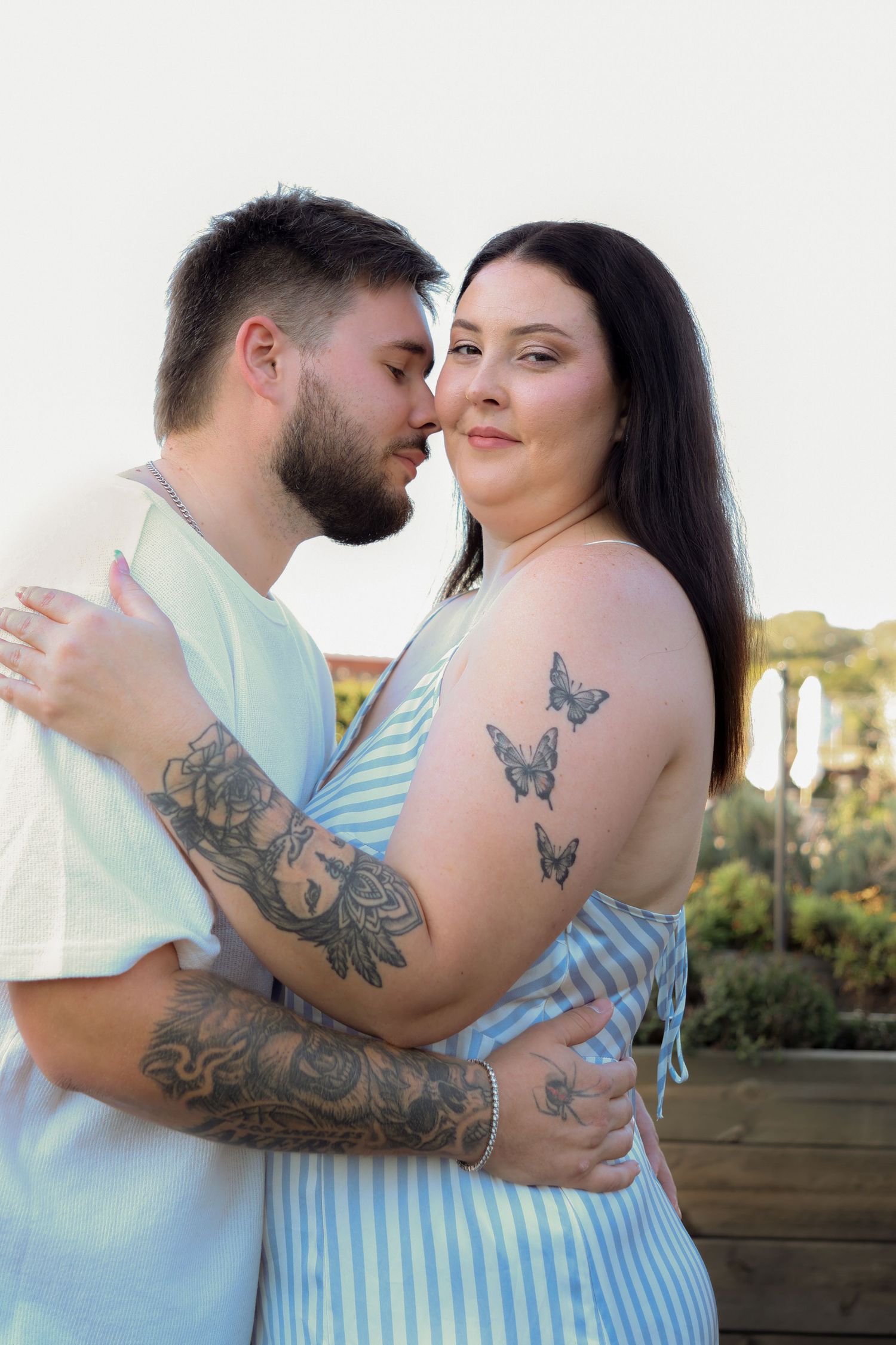 Tattooed couple embracing outdoors, woman with butterfly tattoos in striped dress, man with floral sleeve tattoo.
