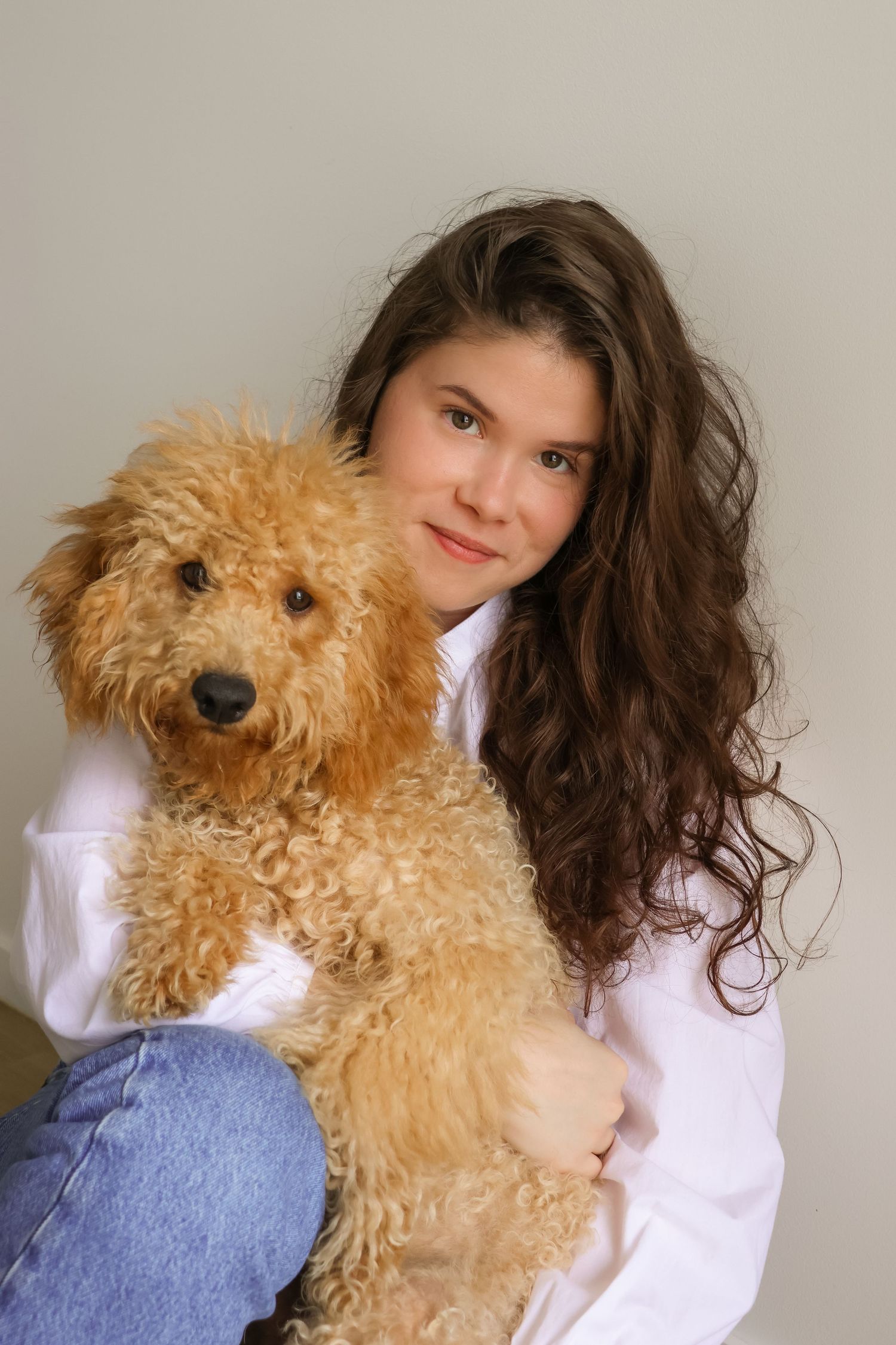 A person in a white shirt and jeans poses with a fluffy goldendoodle dog against a neutral background.
