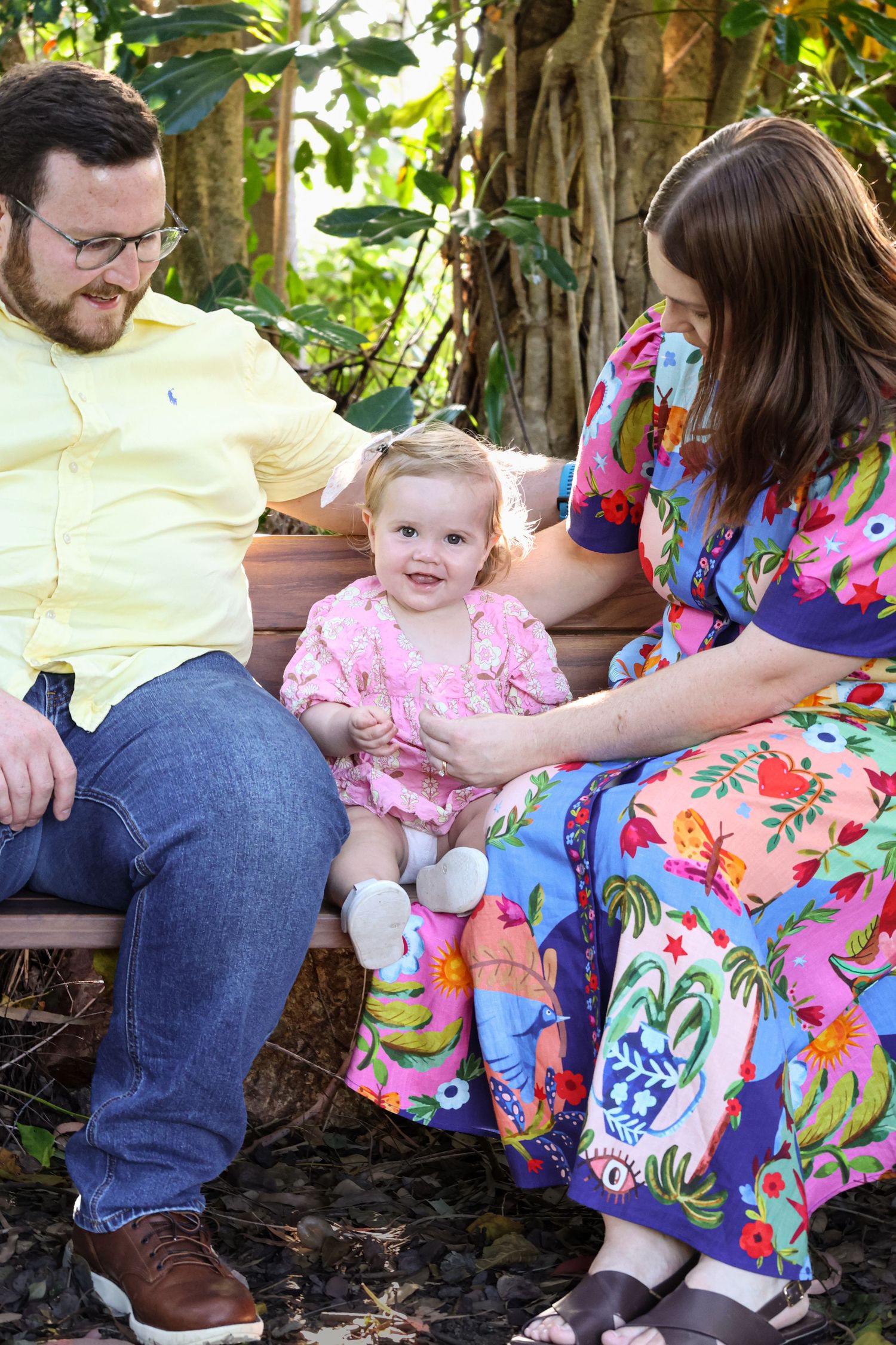 A happy family with a toddler girl in pink dress sits on a bench outdoors, surrounded by lush tropical greenery.