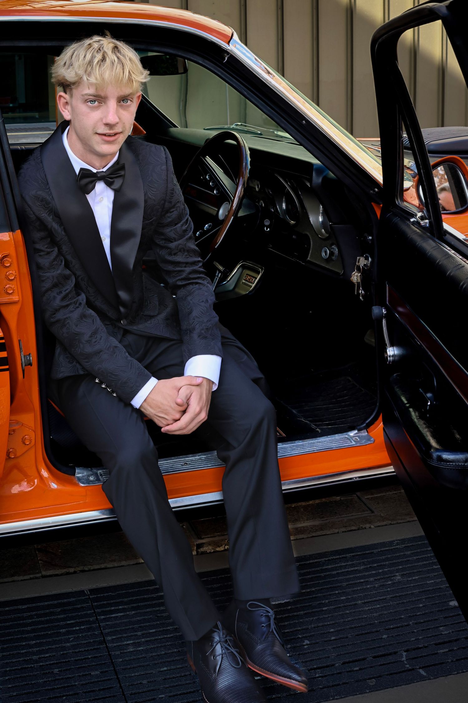 Young man in black tuxedo and bow tie sitting in open door of classic orange car.